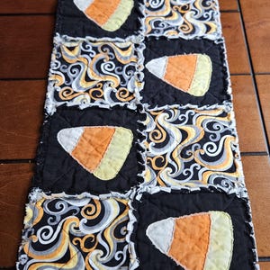 May include: A Halloween-themed table runner featuring alternating black squares with candy corn appliqués and patterned squares with swirls in orange, yellow, gray, and white. The runner is made of fabric with a ragged edge.
