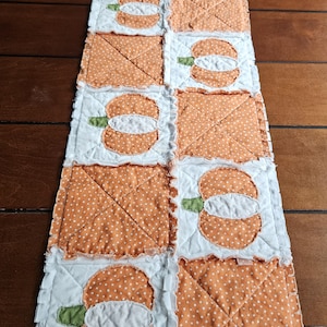 May include: A rectangular rag quilt table runner featuring a repeating pattern of orange and white polka dot fabric squares and white squares with orange pumpkin appliqués. The pumpkin designs include small green stems and visible frayed seams.