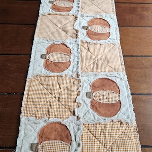 May include: A rectangular rag quilt table runner featuring alternating squares of solid orange pumpkin appliqués and tan and white gingham fabric. The quilt has frayed, exposed seams between each square, creating a textured, layered appearance.