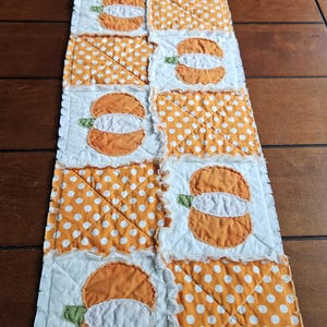 May include: A rectangular rag quilt table runner featuring alternating square patches of orange and white pumpkin appliqués and orange fabric with white polka dots. The quilt has frayed, exposed seams between each square block.
