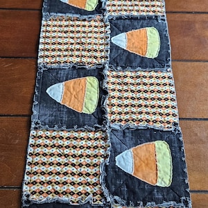 May include: A rectangular rag quilt table runner featuring alternating squares of black fabric with appliqued candy corn shapes and a geometric diamond pattern in orange, black, white, and yellow. The quilt has exposed frayed seams throughout.