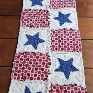 May include: A patriotic table runner featuring alternating blocks of white fabric with blue stars and red, white, and blue patterned fabric. The runner is made of quilted fabric with a frayed edge.