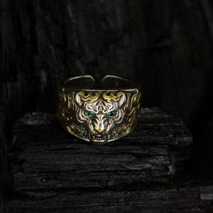May include: A silver-toned ring with a tiger design. The tiger's face is detailed with gold accents and green gemstone eyes. The ring is set against a dark, textured background.