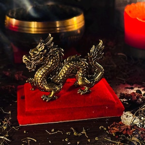 Op de afbeelding: Een gedetailleerd gouden drakenbeeldje met ingewikkelde schubben en kenmerken, geplaatst op een rode fluwelen basis. De draak is het middelpunt, tegen een donkere achtergrond met een kaars en een gouden kom.