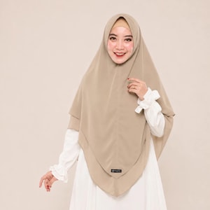 Può includere: Un hijab beige a tinta unita con un design a strati e una piccola etichetta rettangolare nera al centro in basso. Il copricapo è indossato sopra una cuffia interna beige abbinata, insieme a un indumento bianco a maniche lunghe con polsini.