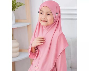 Könnte beinhalten: Ein einfarbiger, rosafarbener Hijab mit glatter Textur, der an beiden Seiten in der Nähe der Schläfen kleine florale Zierelemente aufweist. Die Kopfbedeckung fällt über Schultern und Brust und hat ein kleines rechteckiges Etikett vorne.