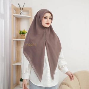Può includere: Un hijab color marrone moka a tinta unita realizzato in un tessuto liscio e leggero che ricade su testa e spalle. Il velo presenta un orlo anteriore pulito e appuntito ed è indossato sopra una camicia bianca a maniche lunghe con il testo anggita.
