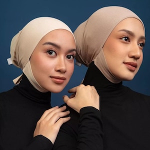 May include: Two beige fabric head coverings worn over black high-neck garments. The head coverings are fitted, smooth, and feature tie-back closures at the nape of the neck, providing full coverage of the hair while framing the face.