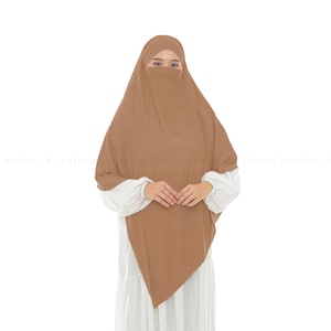 Può includere: Un set khimar e niqab color marrone chiaro a tinta unita, caratterizzato da un lungo copricapo fluido che ricade sulle spalle e sul petto, abbinato a un velo facciale coordinato. Il capo è indossato sopra un abito bianco a maniche lunghe.