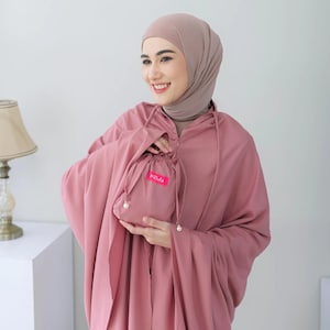 May include: A dusty rose-colored prayer garment featuring a matching hijab and a drawstring pouch. The pouch displays a small, bright pink rectangular label with the white text "Melula" printed on it, accented by two small white beads.