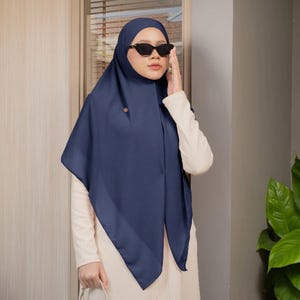 Può includere: Un hijab blu navy a tinta unita con una texture in tessuto liscio e opaco, caratterizzato da una piccola spilla decorativa metallica sul lato. Il velo è drappeggiato in modo semplice, abbinato a un top color crema a maniche lunghe e occhiali neri.