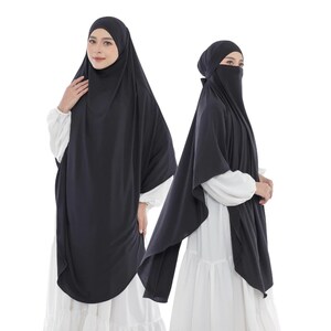 Può includere: Un set composto da hijab e niqab neri indossato sopra un abito bianco a maniche lunghe. Il copricapo nero ricade sulle spalle e sul petto, mentre il niqab abbinato copre la parte inferiore del viso. Il tessuto è nero tinta unita e opaco.