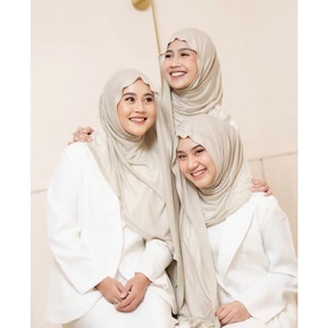 May include: Three individuals wearing matching beige hijabs and crisp white blazers. The hijabs are draped in soft, layered folds around the head and neck, while the white blazers feature structured lapels and long sleeves, creating a coordinated look.