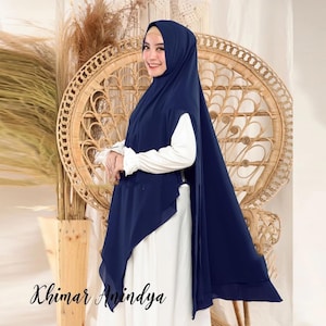 Può includere: Un khimar blu navy con un design a strati fluido che ricade sulle spalle e sulla schiena. Il capo è indossato sopra una maglia bianca a maniche lunghe. Il testo "Khimar Anindya" è stampato in caratteri neri nell'angolo in basso a sinistra.