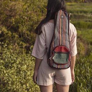 May include: A person wearing a pink dress and carrying a colorful guitar case with a red and green pocket. The case has a woven pattern and is worn over the person's shoulder.