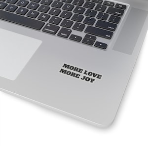 May include: A silver laptop with a black keyboard and a white trackpad. A sticker with the text "MORE LOVE MORE JOY" in a bold, sans-serif font is affixed to the laptop's surface. The image is taken from an elevated angle.