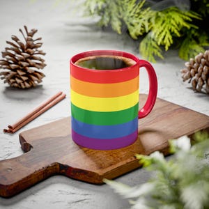 May include: A red-handled coffee mug with a rainbow design, filled with hot coffee. The mug sits on a wooden cutting board, with cinnamon sticks and pine cones nearby. Steam rises from the coffee, suggesting warmth.
