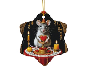 May include: A decorative ornament featuring a gray rat wearing a party hat and holding a heart-shaped object. The rat is positioned in front of a table with candles, an apple, and a cake. The ornament has a gold string for hanging.