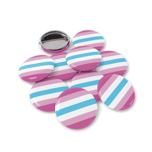 Può includere: Una collezione di bottoni rotondi a righe rosa, bianche e azzurre. Un bottone è capovolto, rivelando un retro in metallo argentato. I bottoni sono sparsi su una superficie bianca.