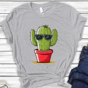 May include: A light gray t-shirt featuring a cartoon cactus wearing sunglasses. The green cactus is in a red pot, set against a yellow background. The shirt is a casual wear item.