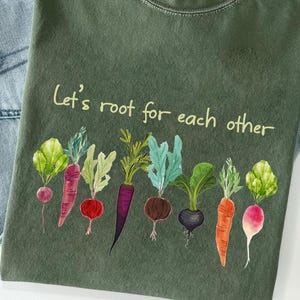 May include: Olive green t-shirt with the phrase "Let's root for each other" and watercolor illustrations of various root vegetables. The vegetables include radishes, carrots, and beets. The shirt is displayed on top of blue denim.