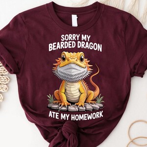 A burgundy t-shirt with a cartoon image of a bearded dragon and text that says "Sorry my bearded dragon ate my homework".