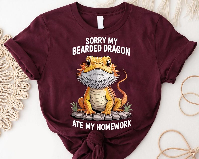A burgundy t-shirt with a cartoon image of a bearded dragon and text that says "Sorry my bearded dragon ate my homework".