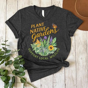 May include: Dark gray t-shirt with the text "PLANT NATIVE GARDENS SUPPORT LOCAL WILDLIFE." The shirt features a colorful floral design with a butterfly. The shirt is knotted at the bottom.
