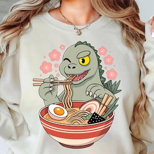 May include: Cream-coloured sweatshirt with a cartoon dinosaur eating ramen. The green dinosaur has a yellow eye and holds chopsticks. The ramen bowl is red and white striped, containing an egg, noodles, and other ingredients.