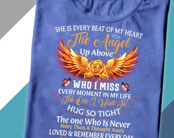 Op de afbeelding: Een blauw t-shirt met de tekst "SHE IS MY MOM". Het shirt heeft een afbeelding van een roos, vleugels en vlinders, met de woorden "The Angel Up Above", "Who I Miss" en "Hug So Tight".