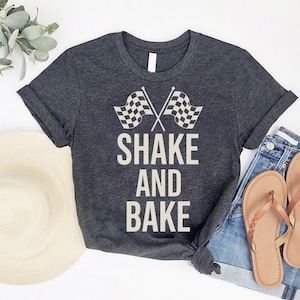 a casual outfit consisting of a gray t-shirt with the text "SHAKE AND BAK" printed on it, a pair of blue jeans, and a pair of brown flip-flops. The outfit is laid out on a white background, with a hat and some greenery visible in the background.