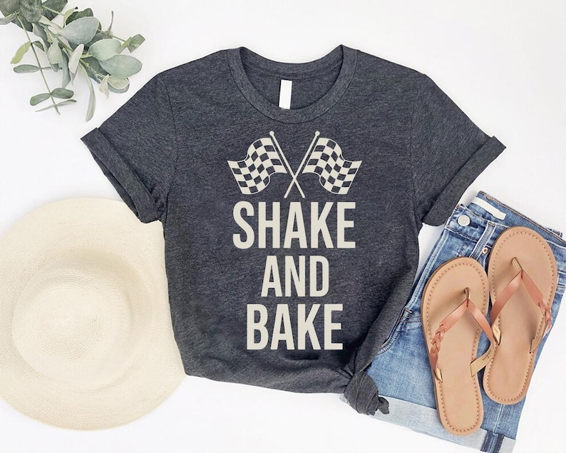 a casual outfit consisting of a gray t-shirt with the text "SHAKE AND BAK" printed on it, a pair of blue jeans, and a pair of brown flip-flops. The outfit is laid out on a white background, with a hat and some greenery visible in the background.