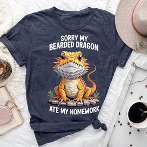 a blue t-shirt with a cartoon image of a bearded dragon and text that says "Sorry my bearded dragon ate my homework". The t-shirt is laid out on a bed, along with a hat, a book, and a cup of coffee.