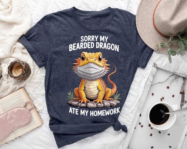 a blue t-shirt with a cartoon image of a bearded dragon and text that says "Sorry my bearded dragon ate my homework". The t-shirt is laid out on a bed, along with a hat, a book, and a cup of coffee.