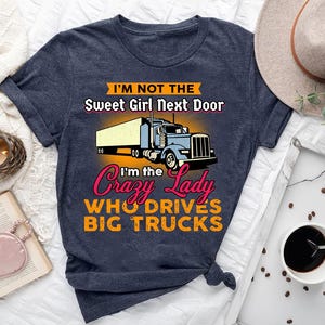 Puede incluir: Camiseta azul marino con el texto "I'M NOT THE Sweet Girl Next Door, I'm the Crazy Lady WHO DRIVES BIG TRUCKS" y un gráfico de un camión azul con un remolque amarillo. La camiseta está anudada en la parte inferior.
