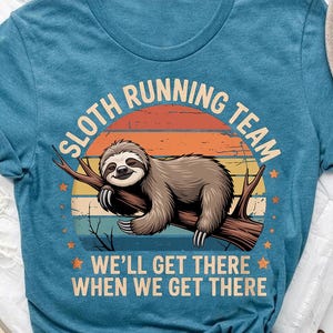 May include: A blue t-shirt featuring a sloth resting on a branch, with the text "SLOTH RUNNING TEAM" above and "WE'LL GET THERE WHEN WE GET THERE" below. The design includes a sunset background and star accents.