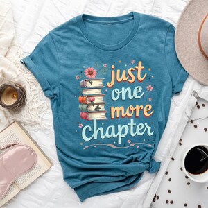 Puede incluir: Camiseta de color turquesa con el texto "just one more chapter" en naranja y azul. El diseño incluye una pila de libros y detalles florales. También se ven un sombrero, café y un libro abierto.