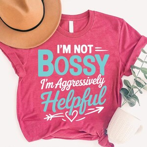 May include: A magenta t-shirt with the text "I'M NOT = BOSSY I'm Aggressively Helpful" in white and turquoise. A brown hat and a white mug are in the background. The shirt has rolled-up sleeves and a knotted bottom.