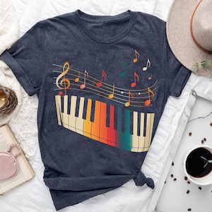 a blue t-shirt with a colorful graphic of a piano keyboard and musical notes, along with a hat, a book, a cup of coffee, and some other items arranged on a bed.