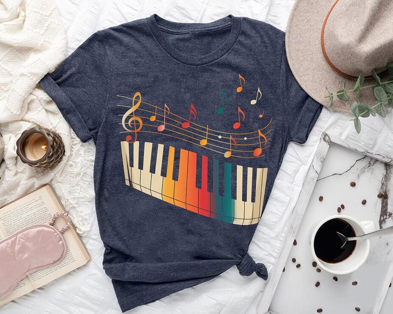 a blue t-shirt with a colorful graphic of a piano keyboard and musical notes, along with a hat, a book, a cup of coffee, and some other items arranged on a bed.