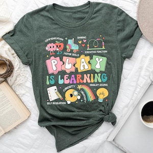 May include: A dark green t-shirt with the words "PLAY IS LEARNING" in colorful letters. The shirt features playful illustrations of a brain, apple, roller skates, and other educational icons. The shirt is knotted at the bottom.