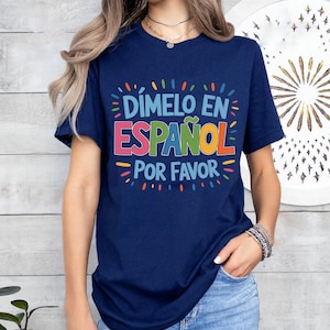 Puede incluir: Camiseta azul marino con el texto "Dímelo en Español por favor" en letras coloridas. El diseño gráfico está en una fuente lúdica con un borde decorativo. La camiseta está hecha de un material suave.