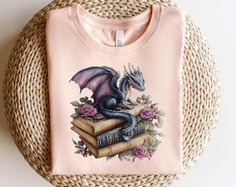 May include: A pink t-shirt with a graphic of a blue dragon perched on top of a stack of old books. The dragon has purple wings and is surrounded by pink roses.