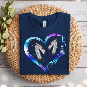 May include: Navy blue t-shirt featuring a heart-shaped design with two feathers and Native American-inspired elements. The heart is outlined in a gradient of blue and purple. The shirt is folded on a woven surface.