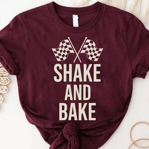 A burgundy t-shirt with the text "SHAKE AND BAKE" printed on it, along with a graphic of a checkered flag.