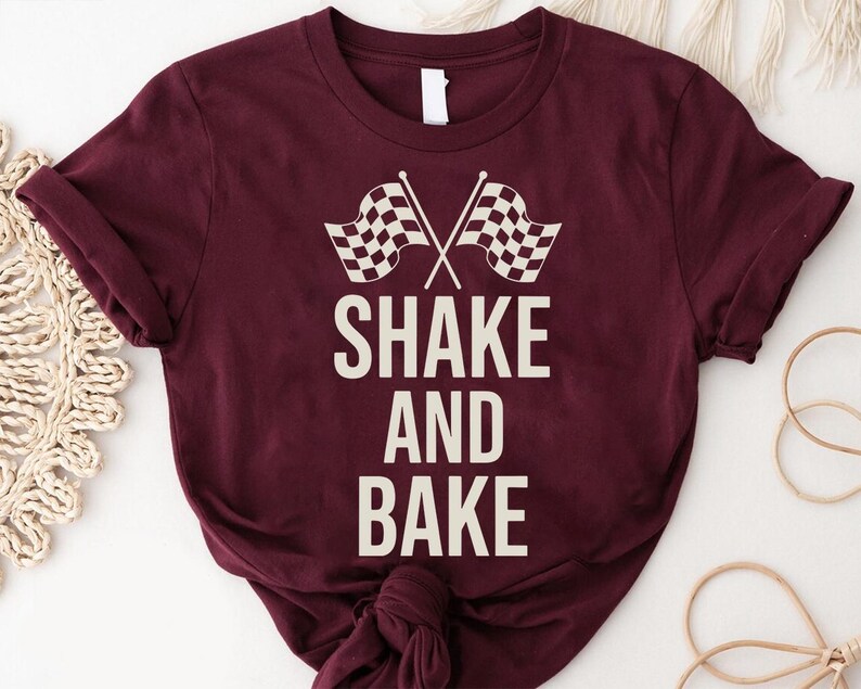 A burgundy t-shirt with the text "SHAKE AND BAKE" printed on it, along with a graphic of a checkered flag.