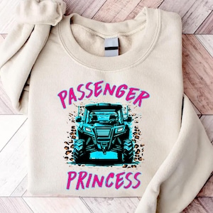 May include: Beige sweatshirt with the words "PASSENGER PRINCESS" in pink and blue. A teal off-road vehicle graphic is centered below the text, with leopard print accents. The sweatshirt is folded on a wooden surface.