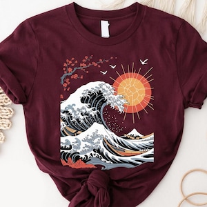 May include: Burgundy t-shirt featuring a Japanese wave design with a rising sun, cherry blossoms, and birds. The graphic is in shades of black, white, orange, and blue. The shirt is made of soft material.