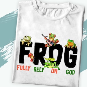 May include: White t-shirt with a black graphic of the word "FROG" with four green frogs in different poses. The text "FULLY RELY ON GOD" is written below the graphic.