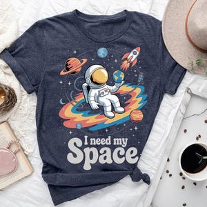 Op de afbeelding: Marineblauw t-shirt met een cartoon astronaut die een wereldbol vasthoudt, omringd door planeten en een raket. De tekst "I need my Space" staat in een retro lettertype. Het ontwerp bevat een kleurrijk regenboogachtig pad.